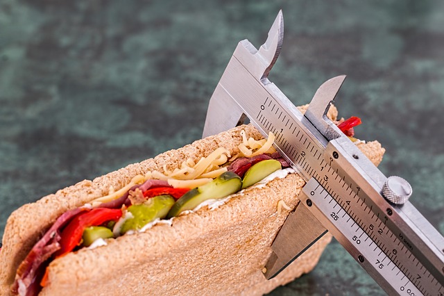 Kovové posuvné meradlo meria hrúbku sendviču. Sendvič je prekrojený a naplnenýuhorkami, červenou paprikou, salámom a nastrúhaným syrom.